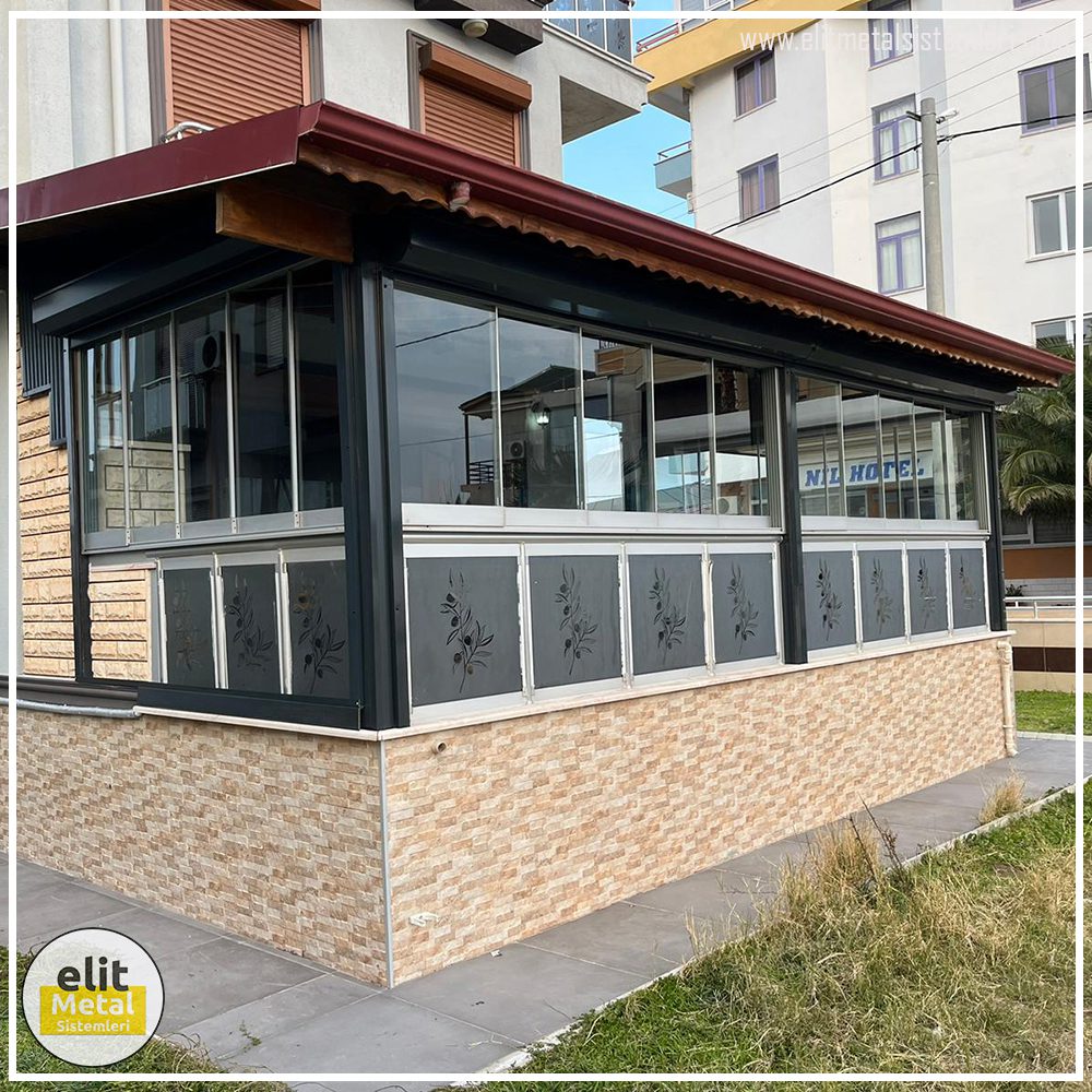 Elit Metal Sistemleri taş duvar üzeri, zeytin dalı motifli dekoratif panelli ve şeffaf camlı teras kapama uygulaması.
